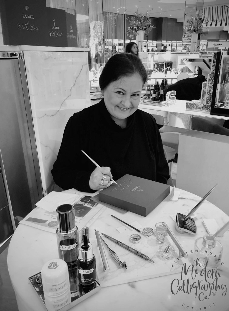 A smiling woman seated at a marble table, engaged in calligraphy while surrounded by beauty products and tools.